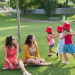 Happy children playing in a sunny park with two mothers sitting nearby, enjoying the outdoors.