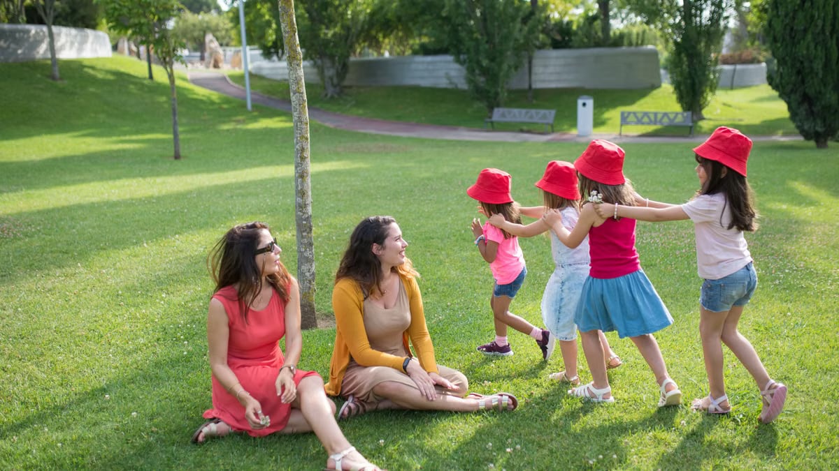 Happy children playing in a sunny park with two mothers sitting nearby, enjoying the outdoors.